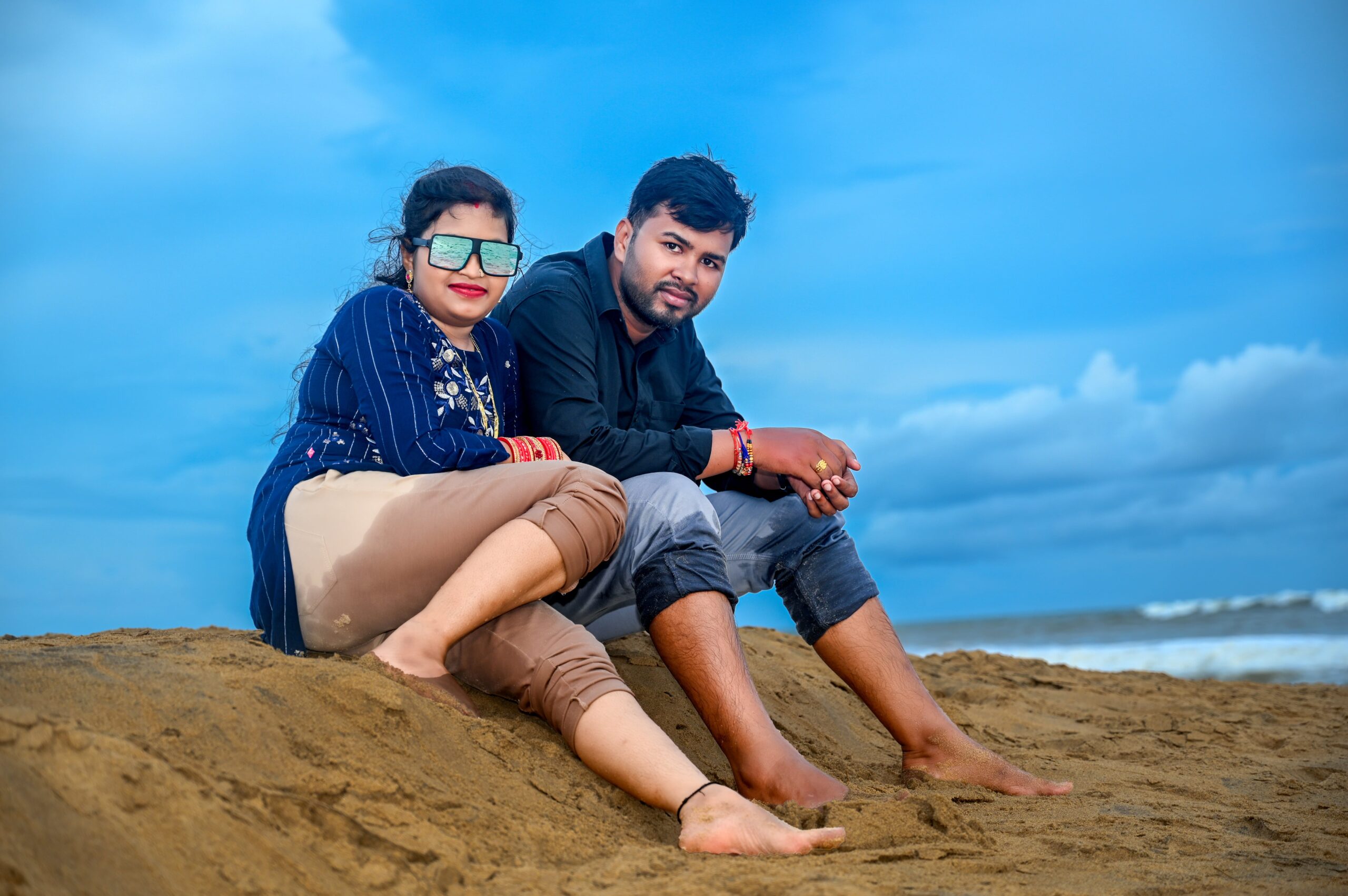Couple sitting barefoot on beach with blue sky and ocean, woman in sunglasses, striped top.