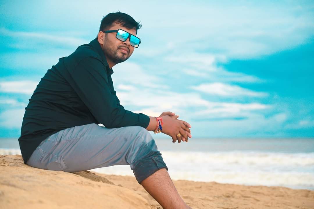 Person in sunglasses, casual clothes, seated on a sandy beach with ocean and cloudy sky backdrop.