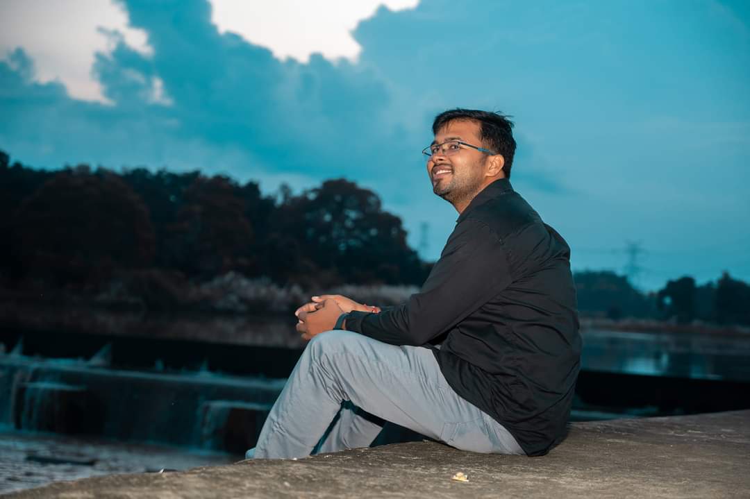 Person sitting on concrete by water, trees, and cloudy sky, wearing glasses, black jacket, gray pants.