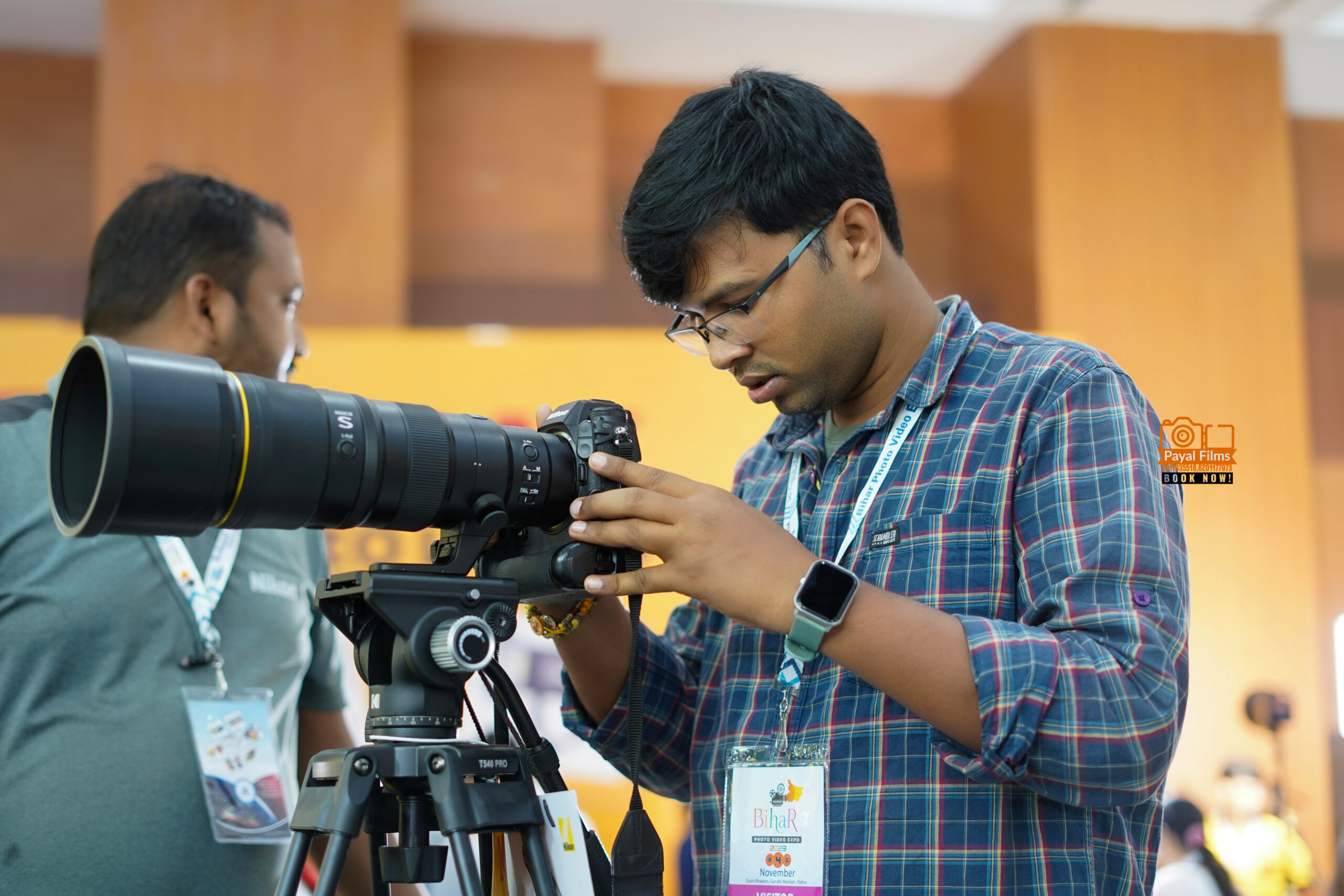 Payal Films owner adjusting professional Nikon camera on tripod during a photography event in Bihar, wearing checkered shirt and event badge