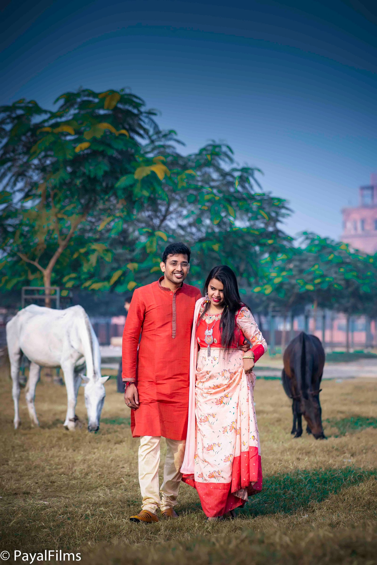 Indian couple in red kurta and peach floral dress posing outdoors with horses by Payal Films