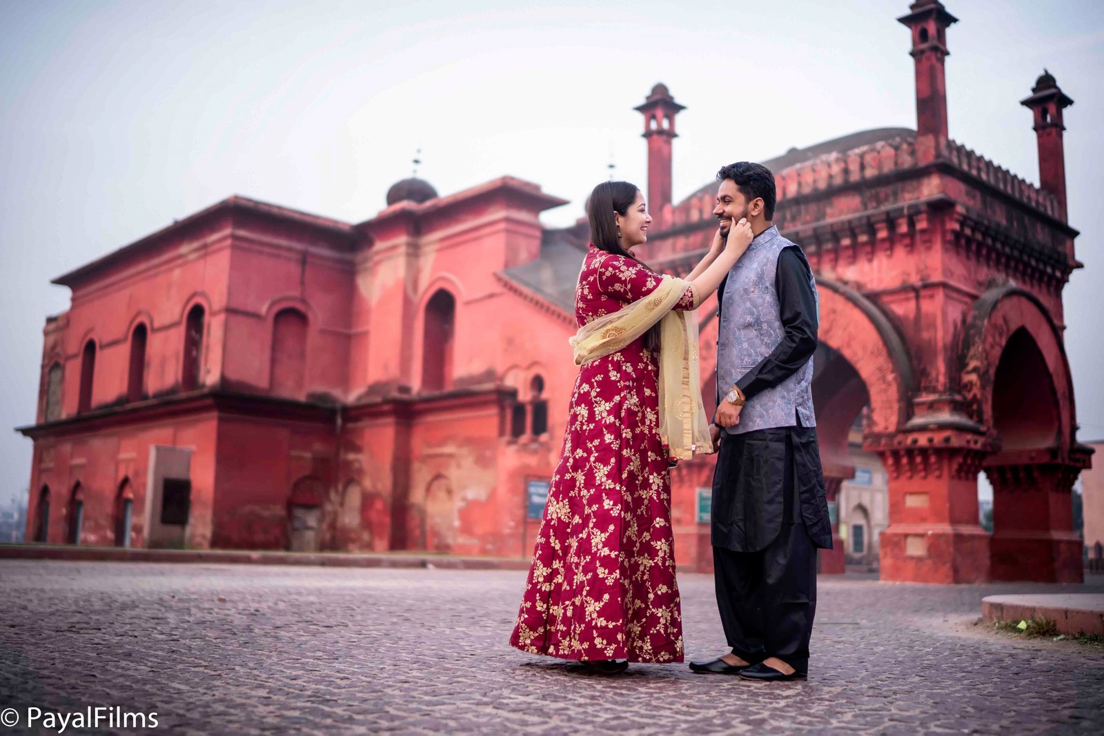 A couple in traditional Indian attire posing back-to-back in front of a historic monument by Payal Films.
