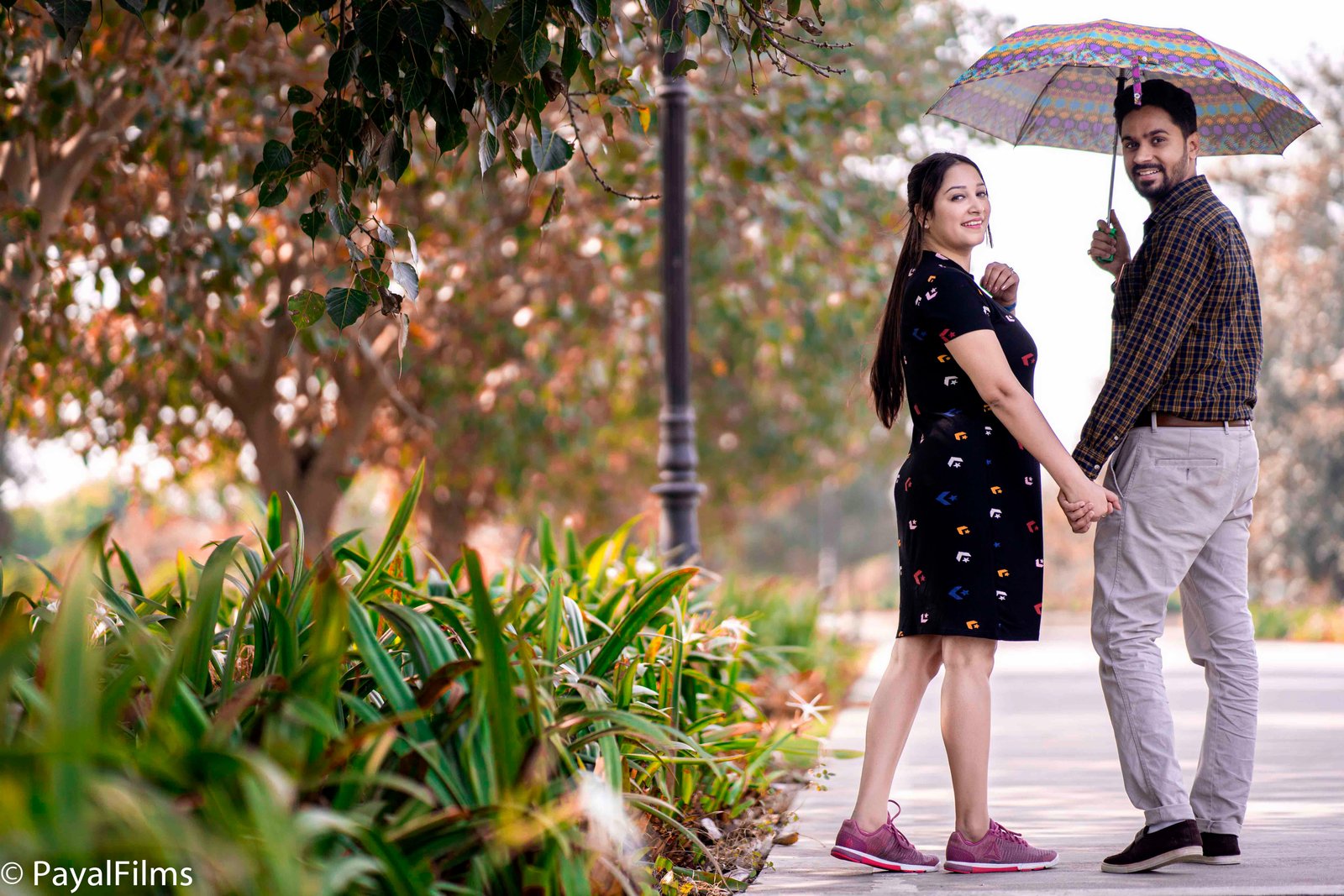 Romantic couple holding hands and smiling under a colorful umbrella in a park by Payal Films