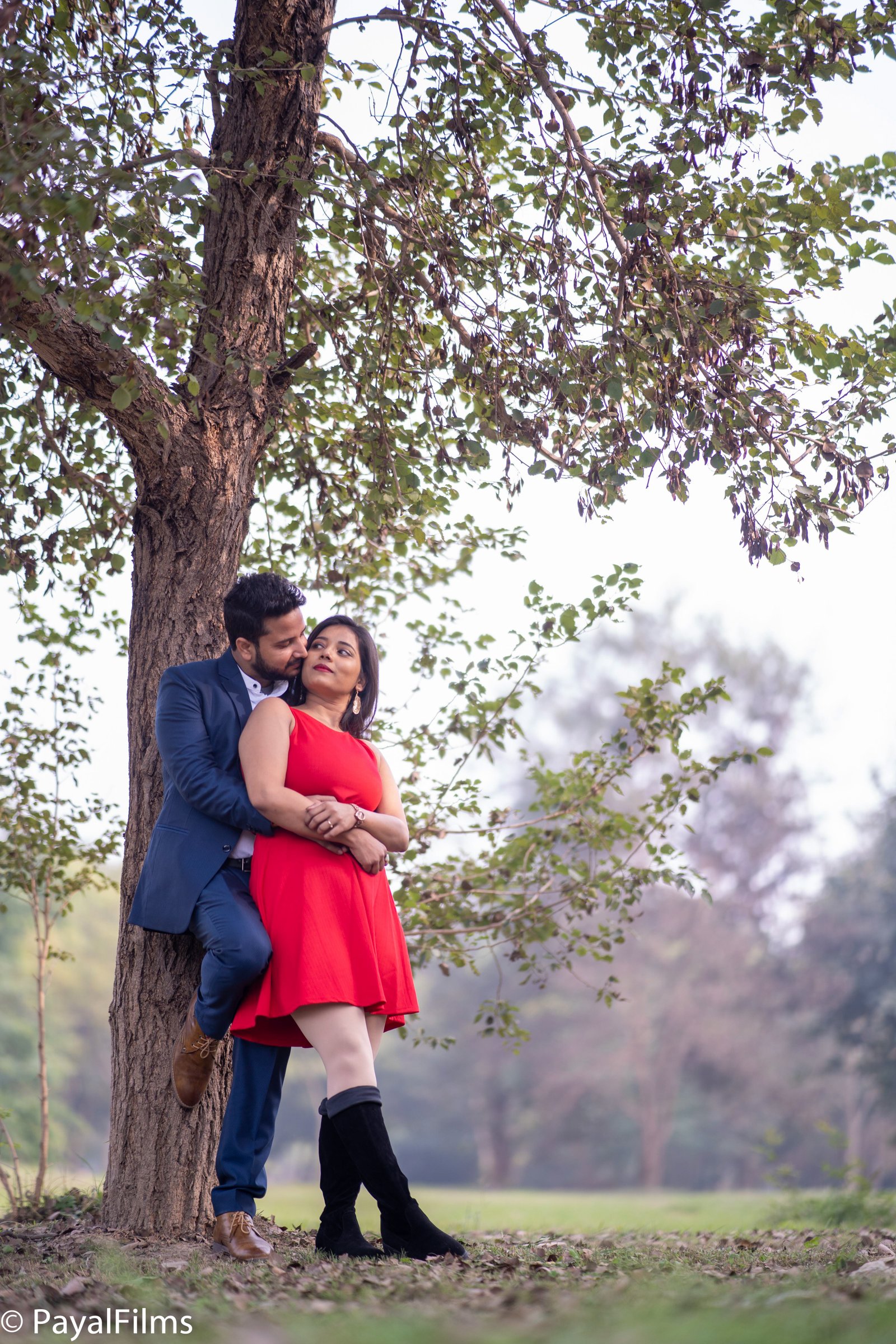 Romantic pre-wedding couple hugging by a tree, woman in red dress and man in blue suit by Payal Films.