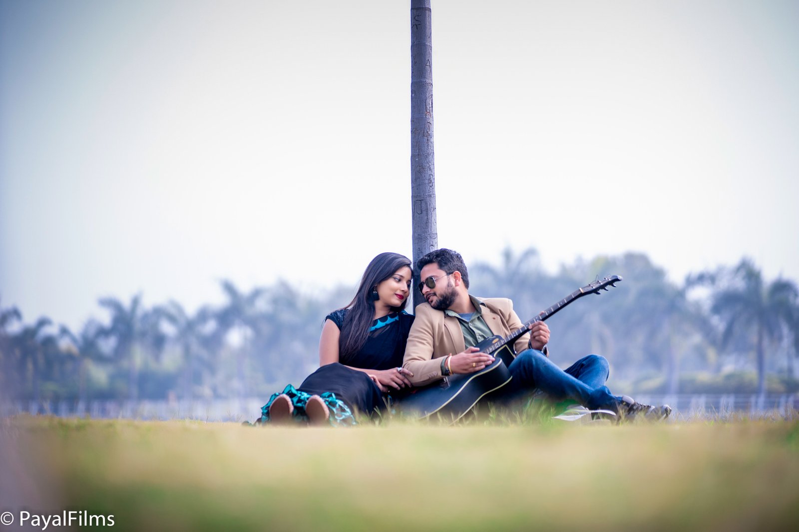 Romantic couple sitting together outdoors leaning against a pole while playing guitar, captured by Payal Films.