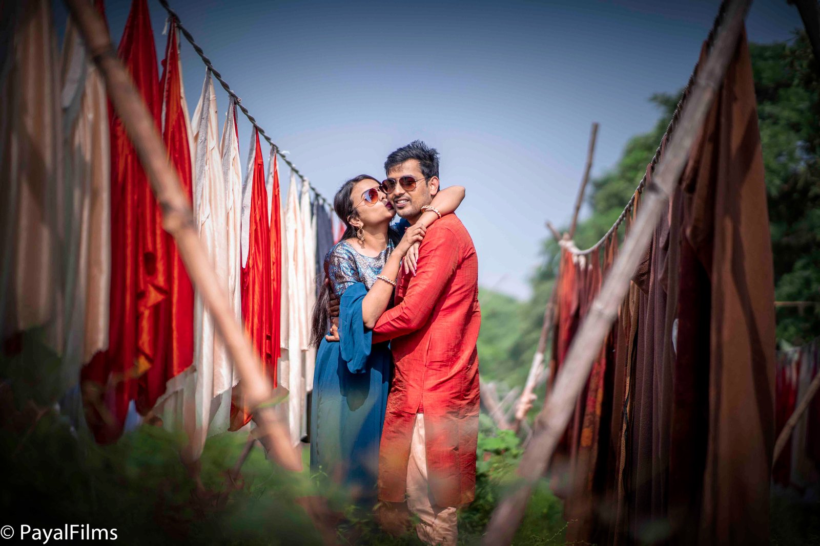 Romantic outdoor pre-wedding couple portrait with woman in blue dress kissing man in red kurta by Payal Films