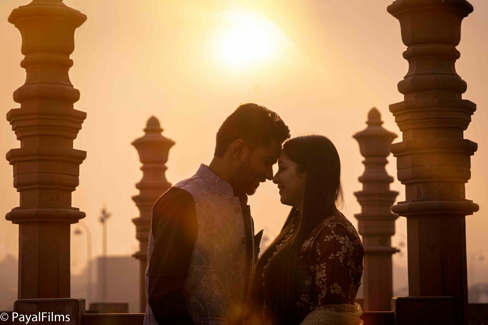 A couple in traditional maroon attire holding hands on red stone stairs, captured by Payal Films in a cinematic style.