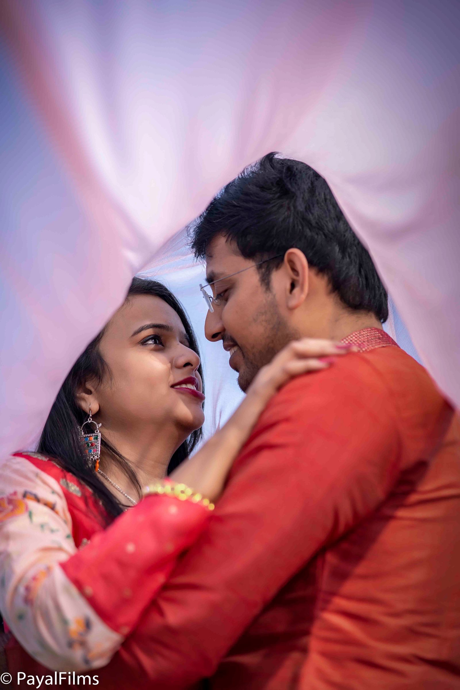 Romantic Indian couple in traditional attire looking lovingly at each other under a pink veil captured by Payal Films.