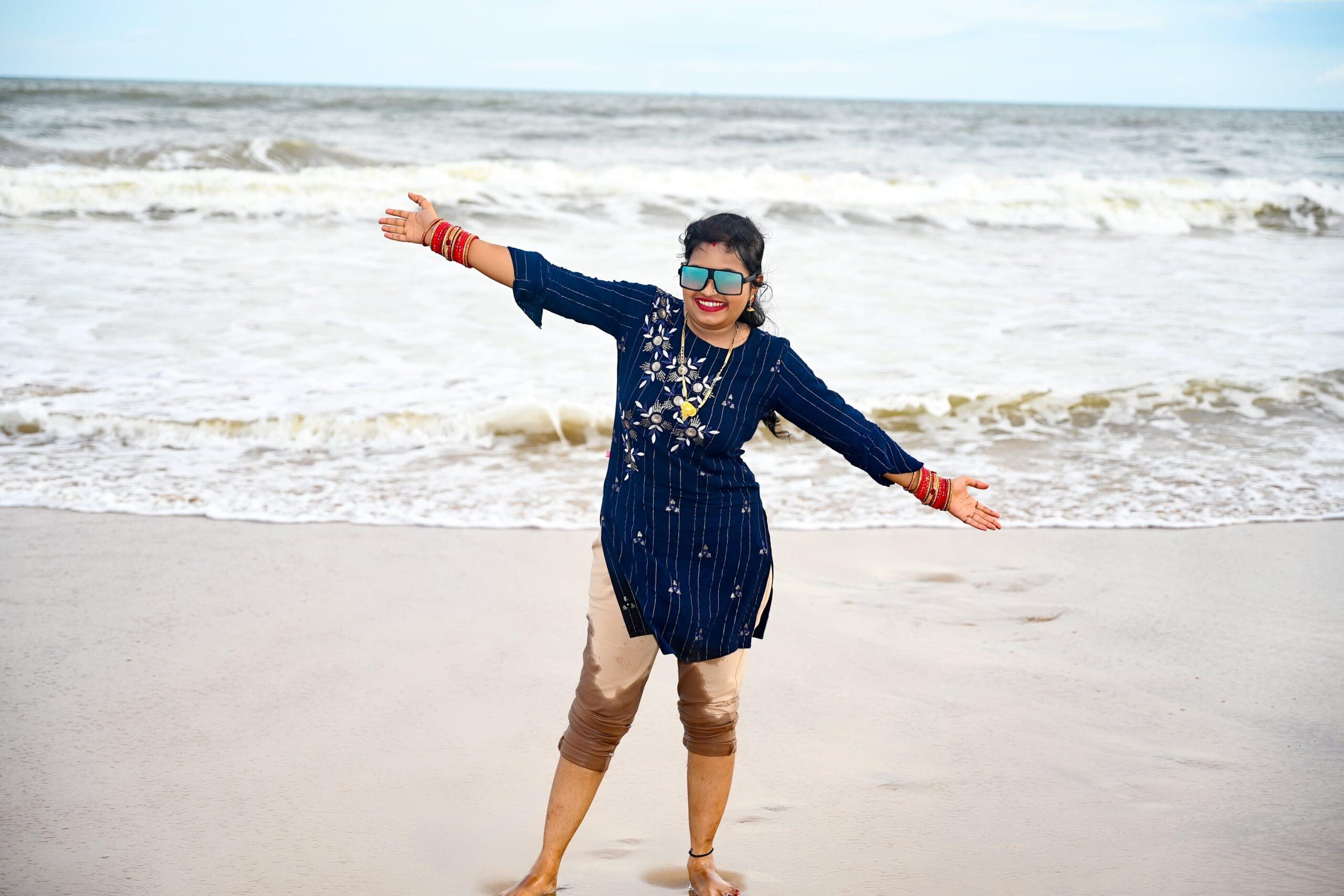 Girl joyfully poses on sandy beach in blue tunic and capri pants with ocean in background.