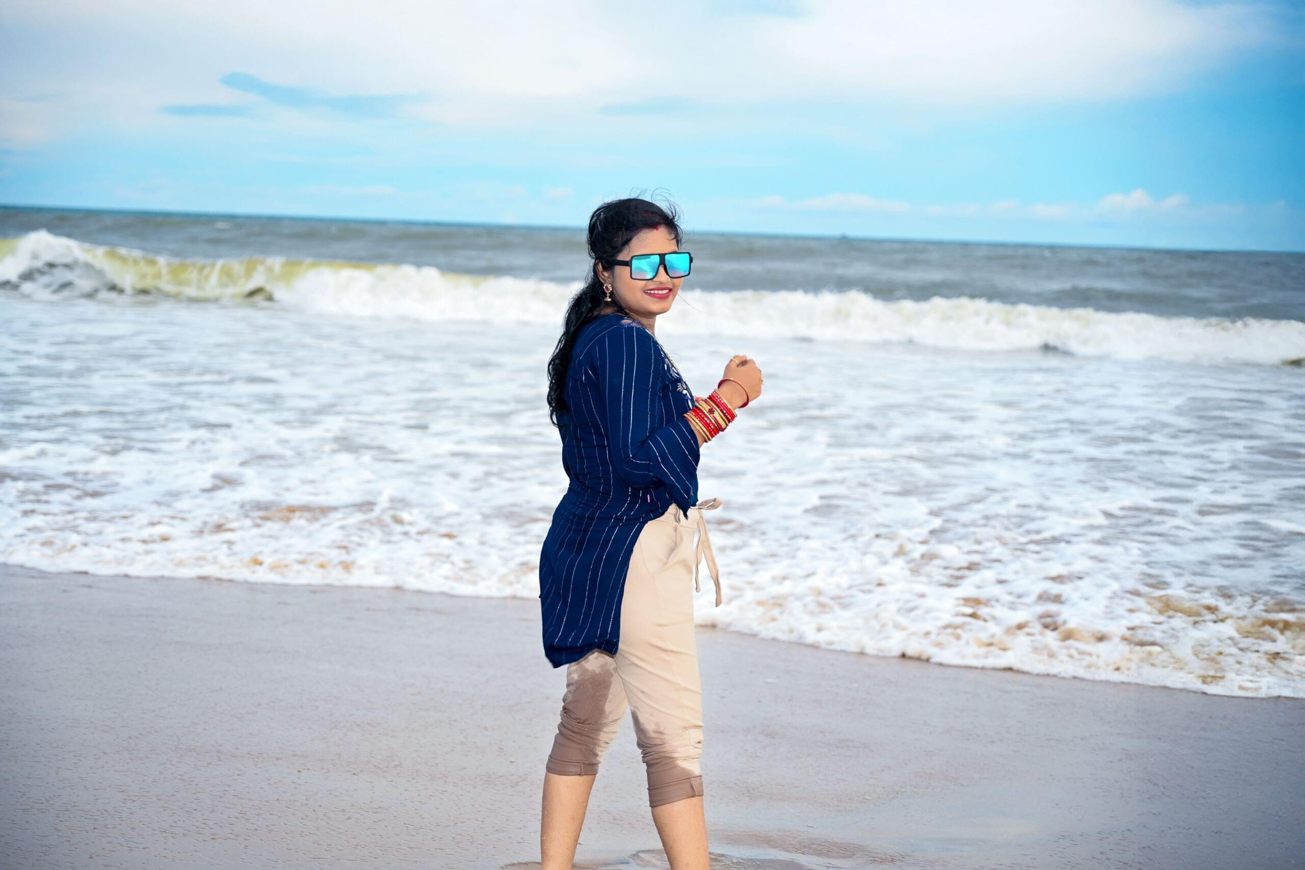 Person in sunglasses and striped blue top smiling on a sandy beach with waves in background.