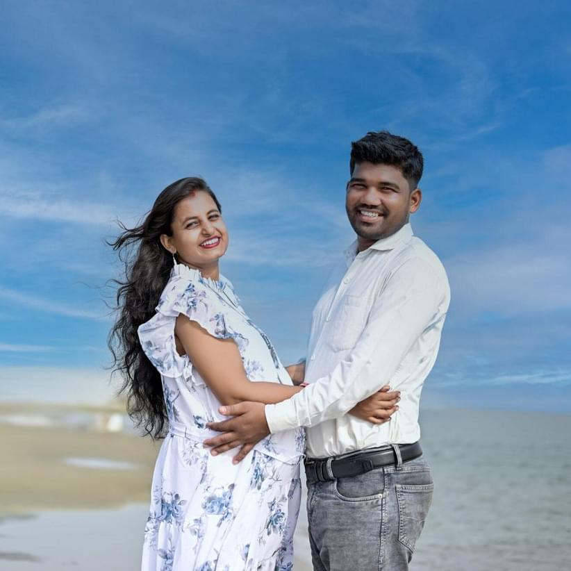 A happy couple embraces during a maternity photoshoot at the beach. The woman, wearing a white floral dress, smiles as her partner holds her close under a beautiful blue sky.