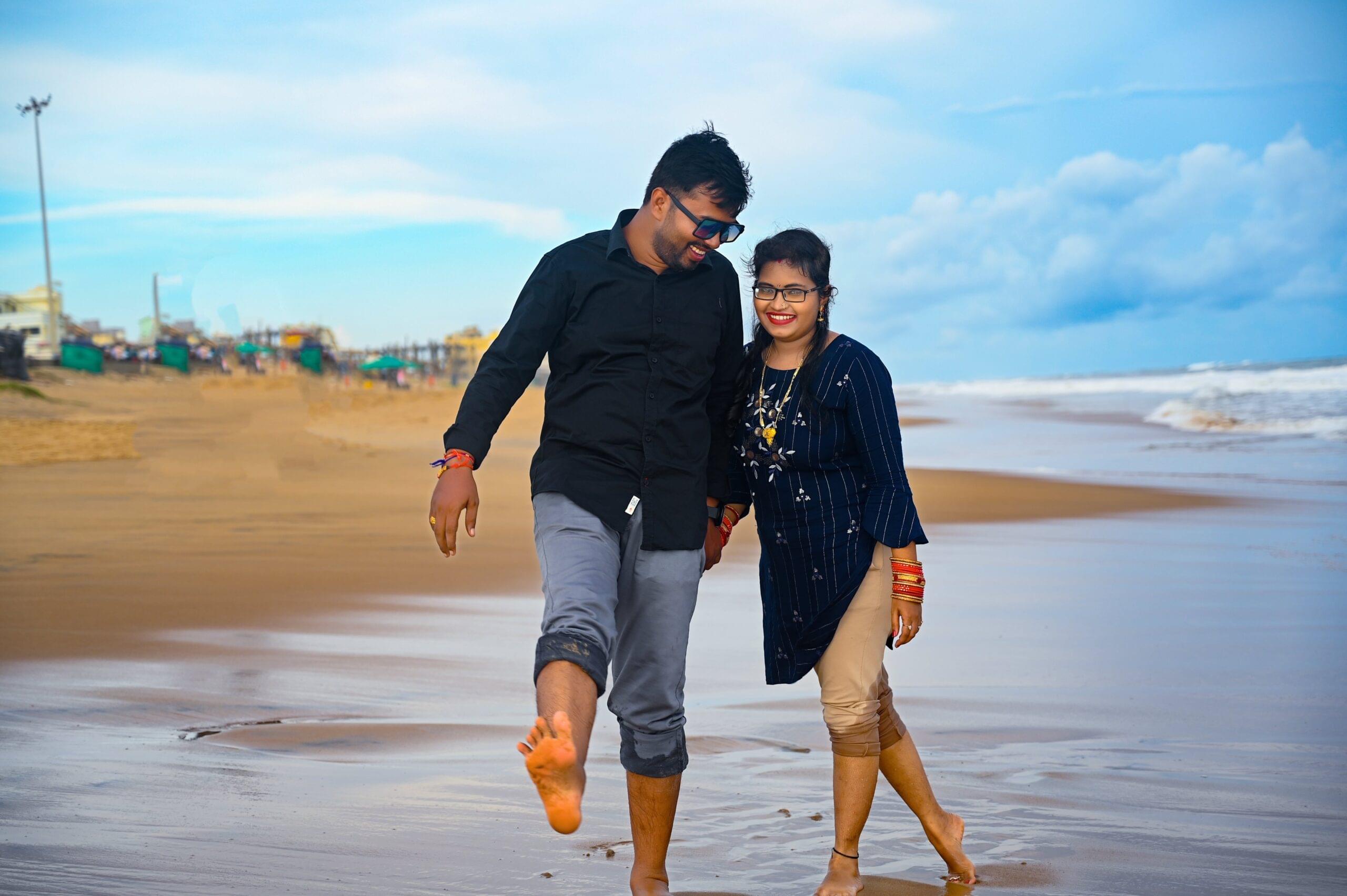 Couple walking barefoot on sandy beach, man lifting foot playfully, shoreline in background.