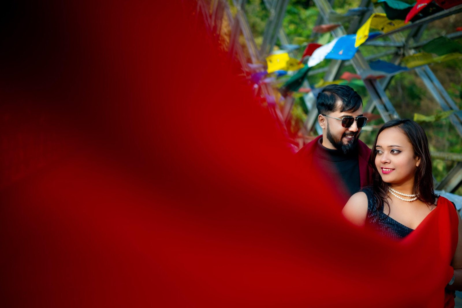 Couple smiling outdoors near colorful flags, partially obscured by a red fabric in the foreground - Payal Films Bihar