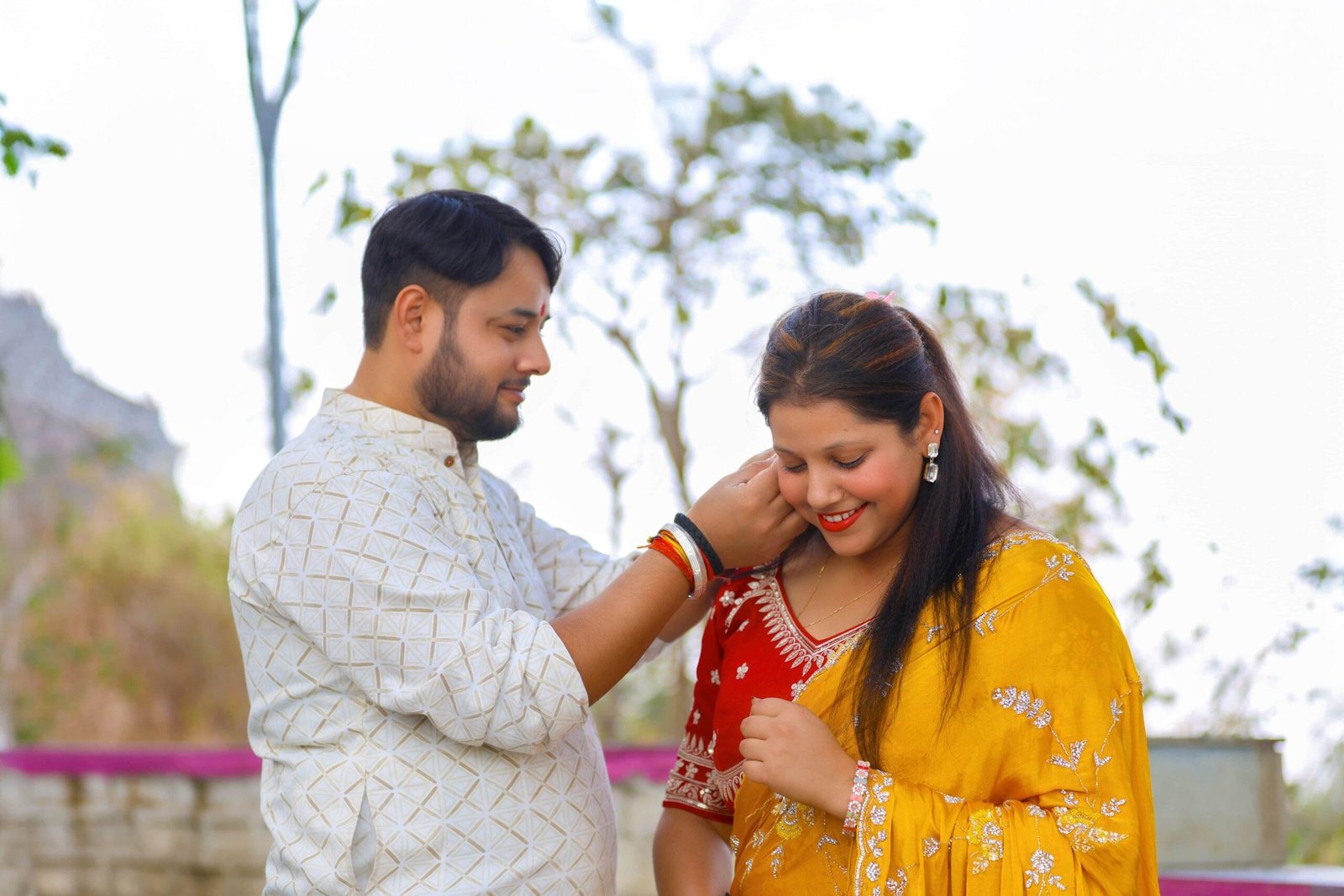 Man adjusting woman's earring outdoors; she smiles in a yellow saree; trees in background - Payal Films Bihar