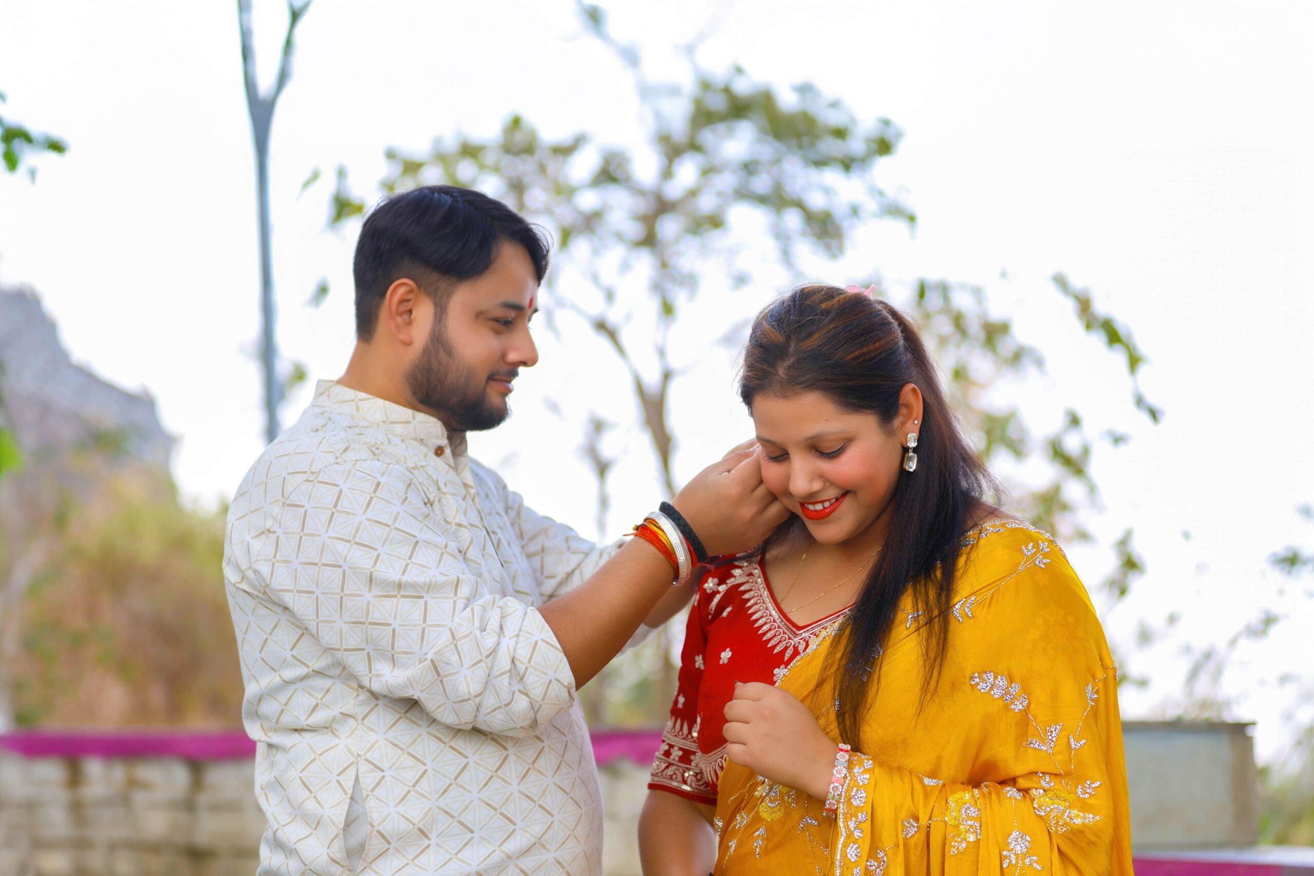 Man adjusting woman's earring outdoors; she smiles in a yellow saree; trees in background.
