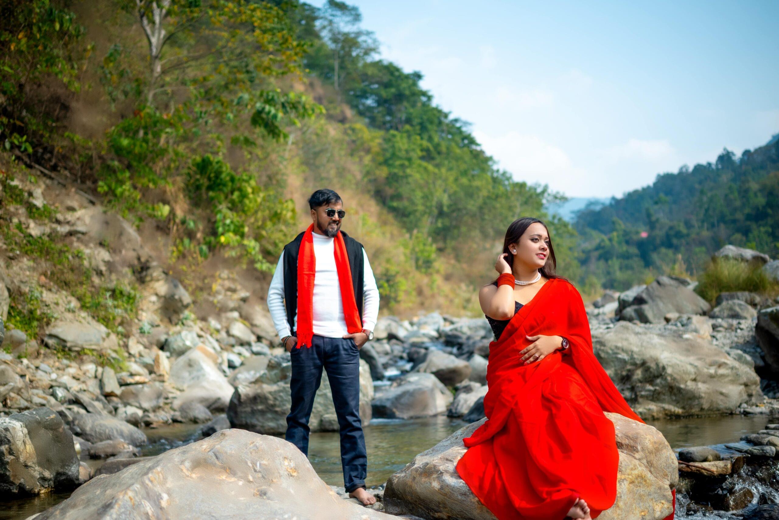 Man and woman in vibrant outfits outdoors, with rocks, stream, and forested hills in background.