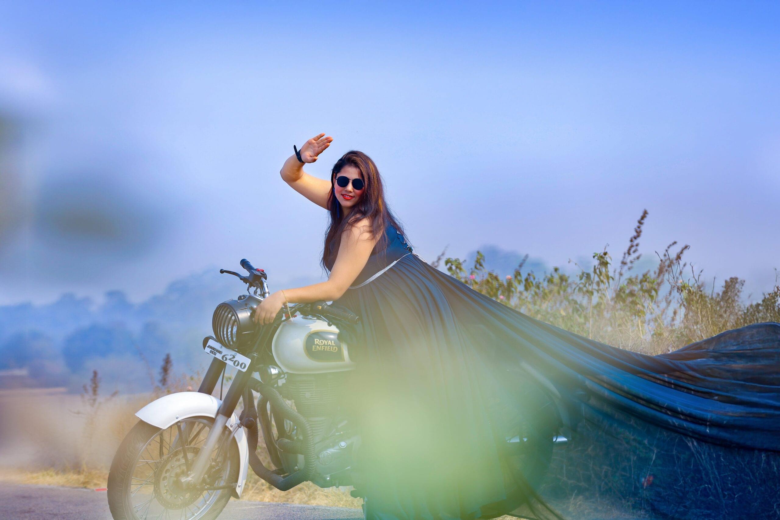 Person in sunglasses sitting on Royal Enfield motorcycle with long outfit outdoors.