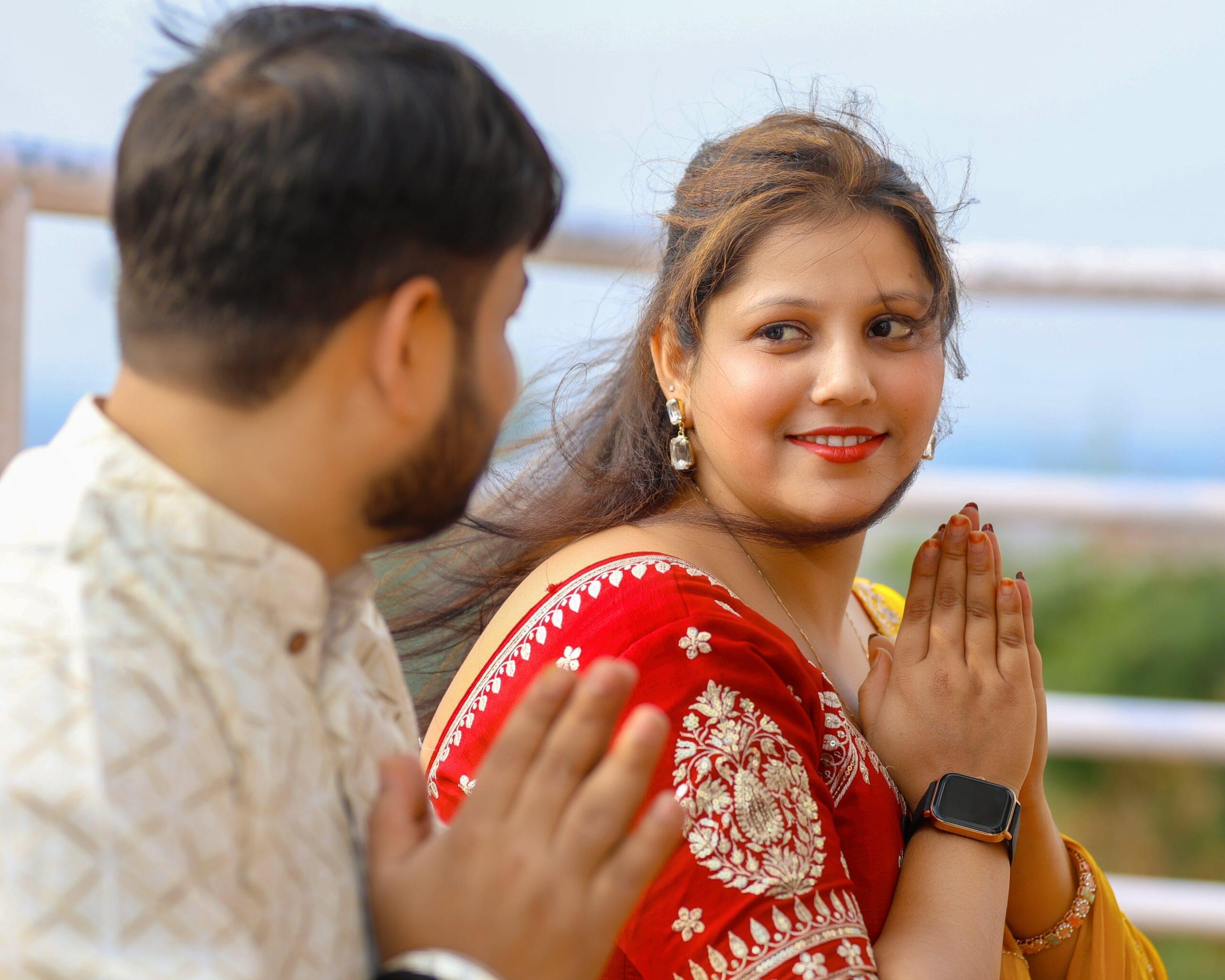 Man and woman outdoors, palms together in traditional greeting, woman in red outfit with smartwatch.