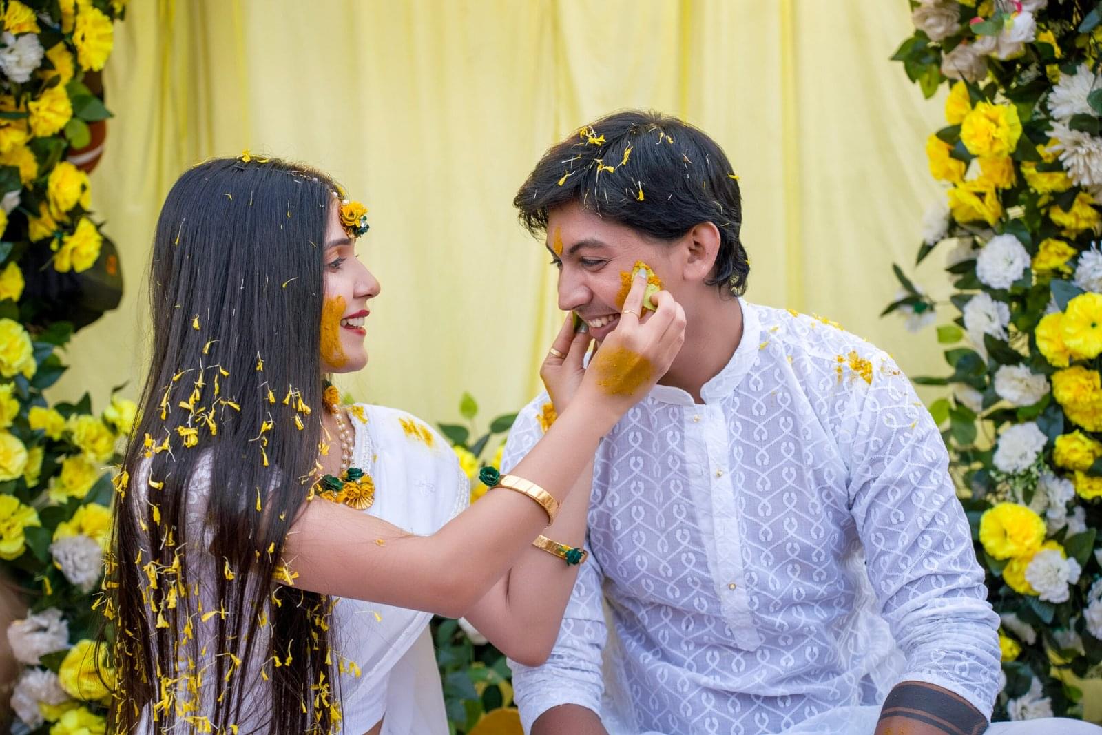 Two people in white apply turmeric paste, smiling amid yellow, white flowers in a festive setting.
