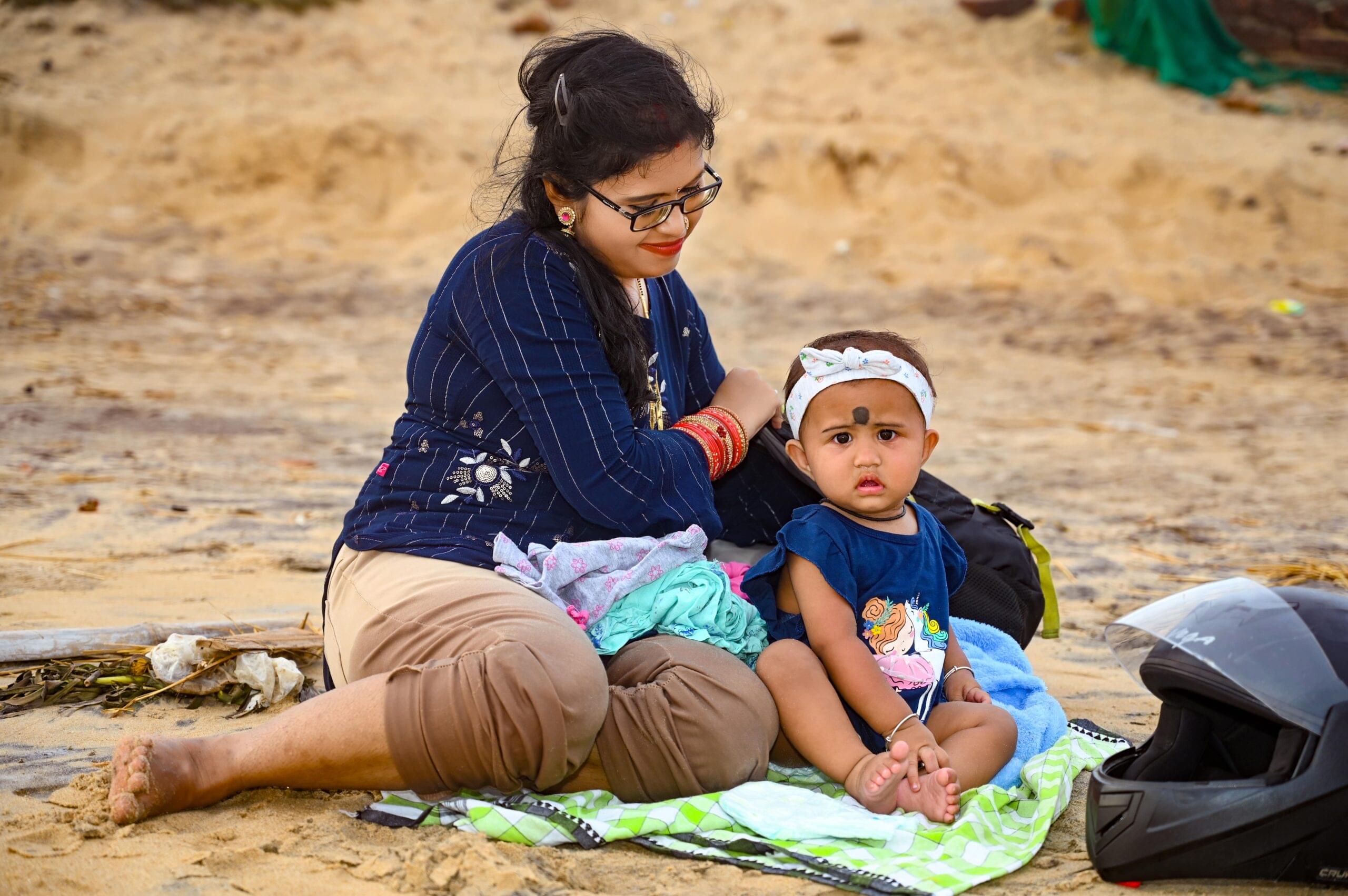 Woman and baby on beach with helmet, sitting on a mat with scattered clothing items.