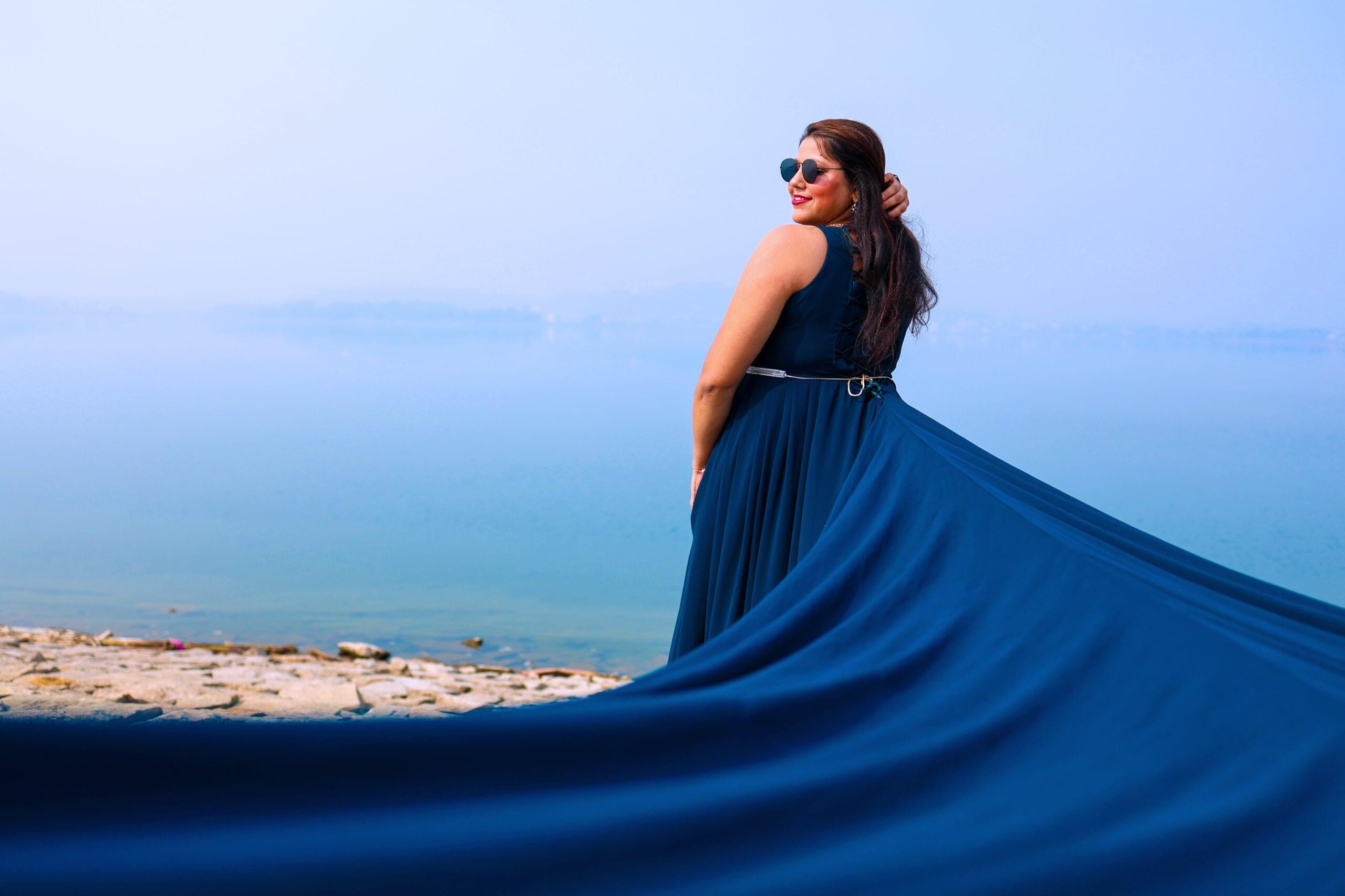 Woman in blue dress and sunglasses by calm water with flowing dress behind her.