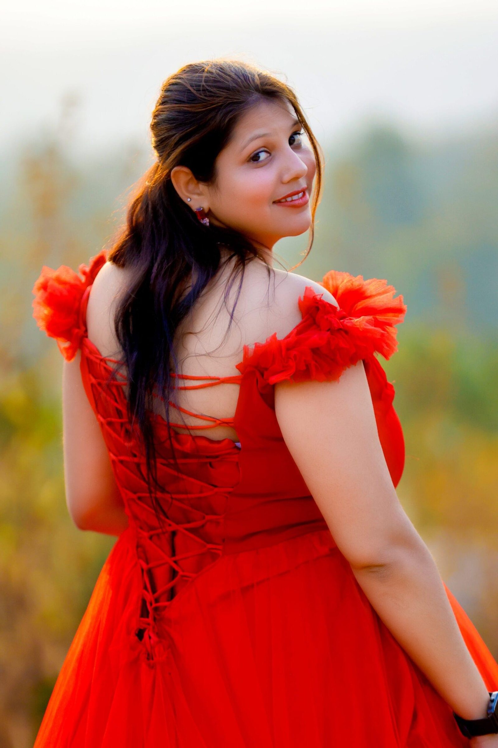 Woman in red off-shoulder dress with back lace-up in natural setting, looking over shoulder - Payal Films Bihar