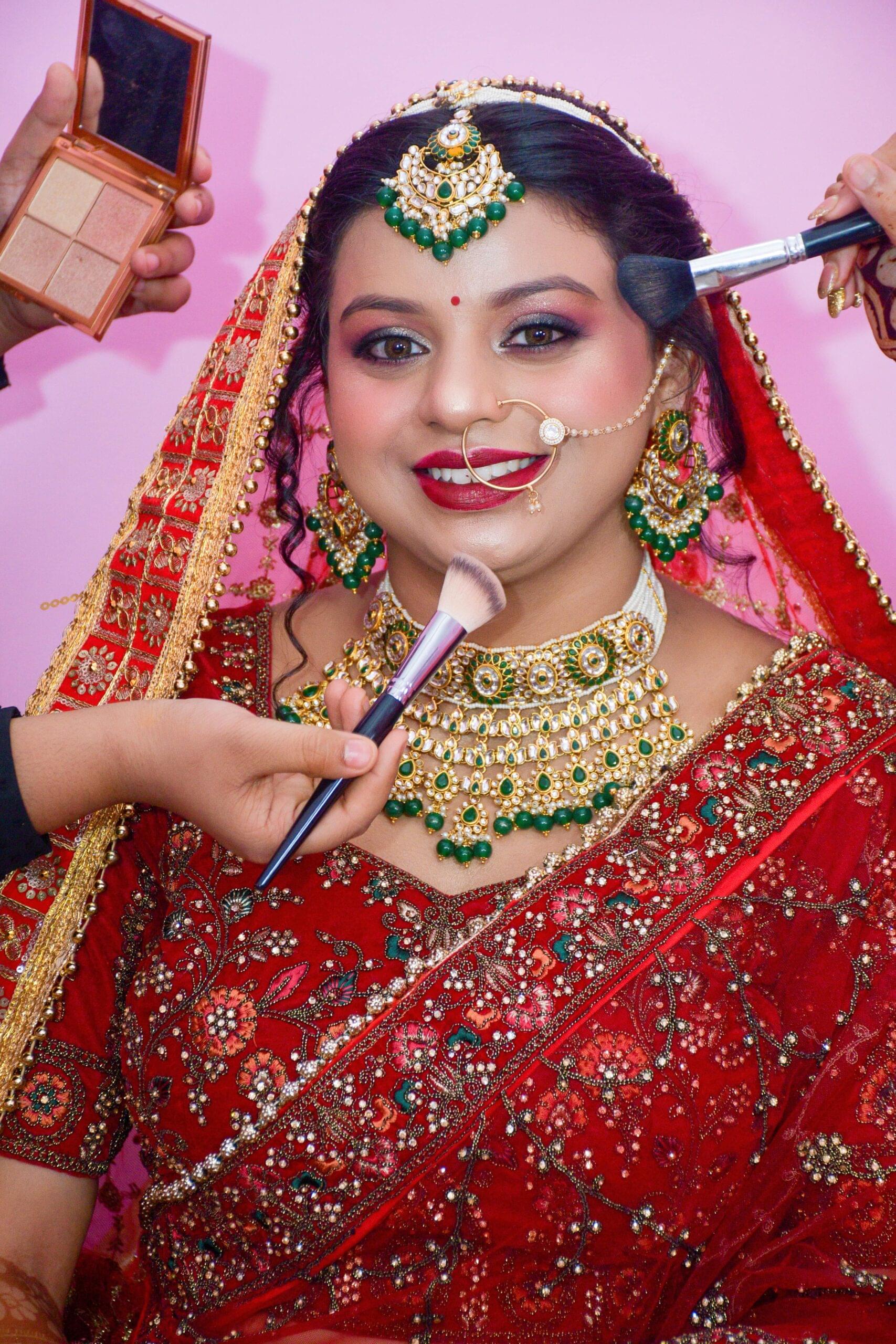 Woman in red and gold sari with jewelry, having makeup applied for an occasion.