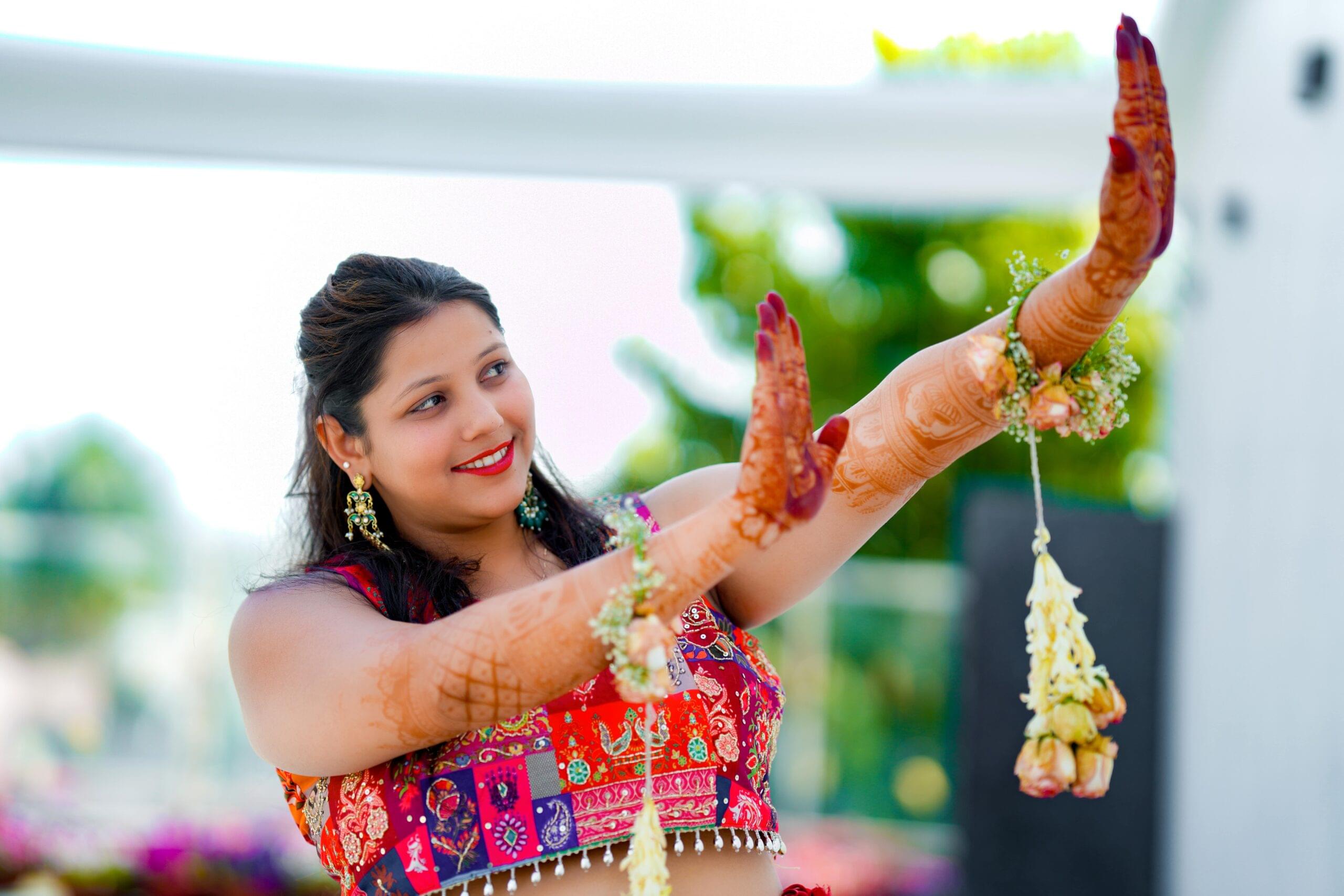 Woman in vibrant traditional attire with henna and floral jewelry, joyfully posing outdoors.