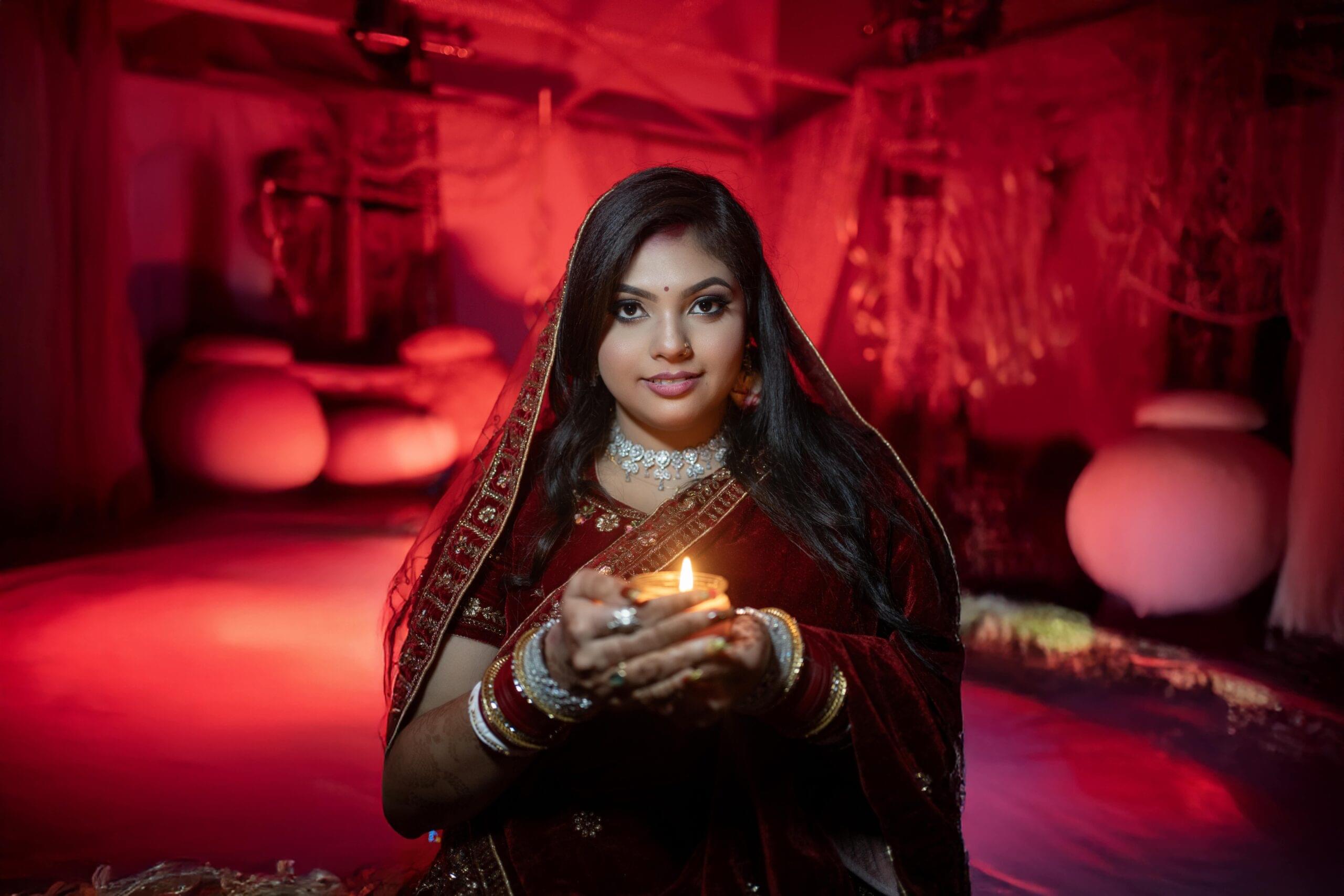 A stunning Indian bride in a deep red bridal lehenga, holding a glowing diya, captured by Payal Films Photography in Khagaria, Bihar. The warm ambiance and traditional setting enhance the beauty of this cultural wedding portrait.