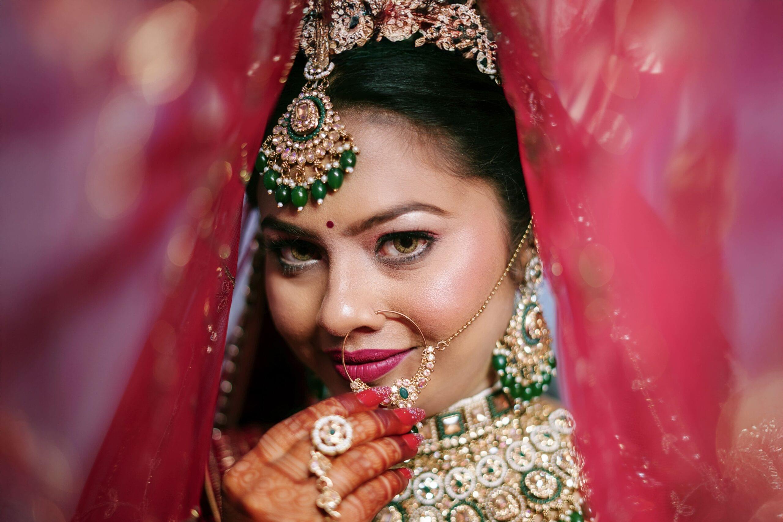 Closeup portrait of an Indian bride seen through a red veil, showing detailed jewelry, mehndi and bridal dress